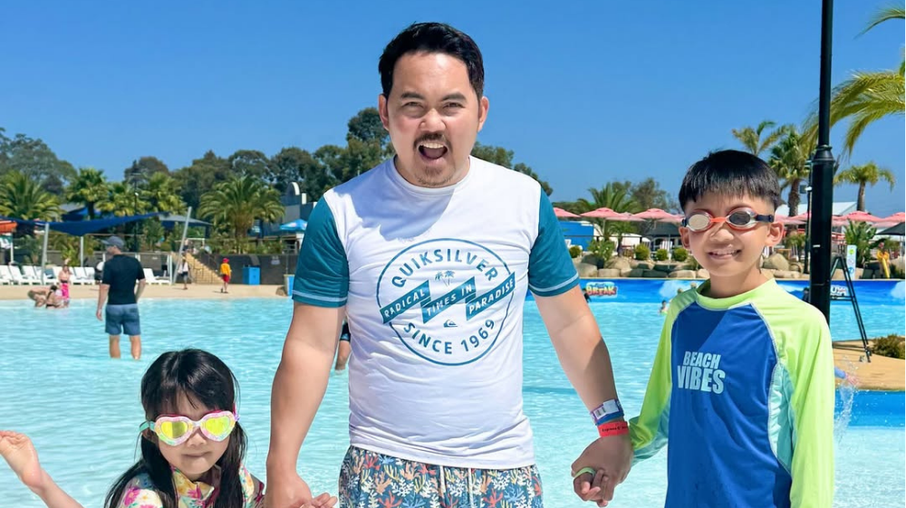A father and his two children smile and pose in front of the wave pool at Gumbuya World on a sunny day. All wear swimwear and goggles, capturing a fun family moment from the influencer campaign highlighting the park's family-friendly experience.