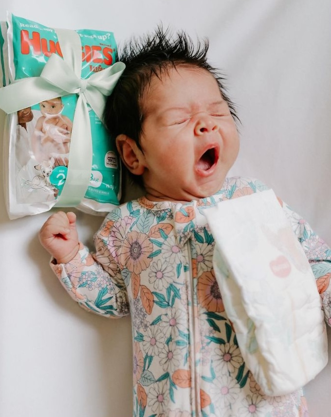 A yawning newborn baby wearing a floral onesie lies on a white background next to a ribbon-wrapped pack of Huggies Newborn Zinc nappies. A single nappy is resting on the baby’s chest, highlighting the product in a sweet and candid moment.