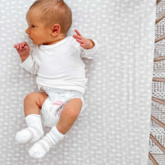 A newborn baby wearing a white long-sleeve onesie, socks, and a Huggies nappy lies on a light grey patterned sheet inside a cot. The baby is looking to the side with arms gently raised. Social Soup Networker and Micro Creator Campaign.