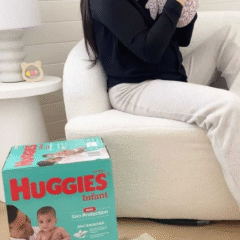 A smiling mother sits on a cream armchair holding her baby up in a playful moment. In the foreground, a box of Huggies Infant Zinc-Enriched Nappies (Size 2, 4–8kg) is placed on a cream rug, clearly showing the product branding. Social Soup Networker and Micro Creator Campaign.