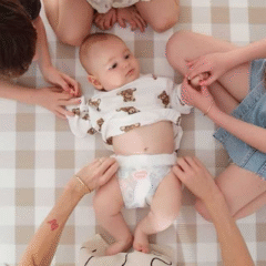 A baby wearing a printed top and Huggies nappy lies on a checkered mat, surrounded by siblings and a parent gently holding their hands and legs. The scene captures a warm, caring family moment from a top-down perspective. Social Soup Networker and Micro Creator Campaign.