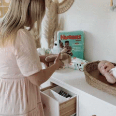 A mother stands beside a white dresser preparing baby items while her smiling baby lies in a woven changing basket. A pack of Huggies Newborn Zinc nappies is visible on the dresser, adding context to the calm and cosy nursery setting. Social Soup Networker and Micro Creator Campaign.